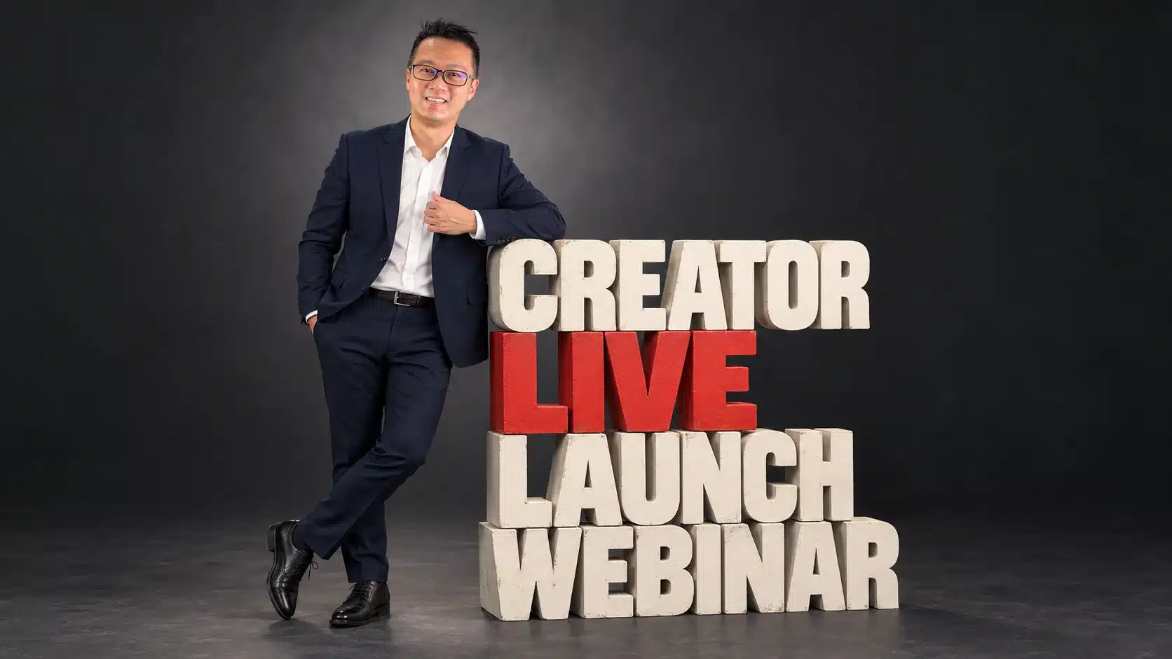 Terence Tam in a navy suit leaning on large stone block letters spelling “Creator Live Launch Webinar,” with “Live” in red.